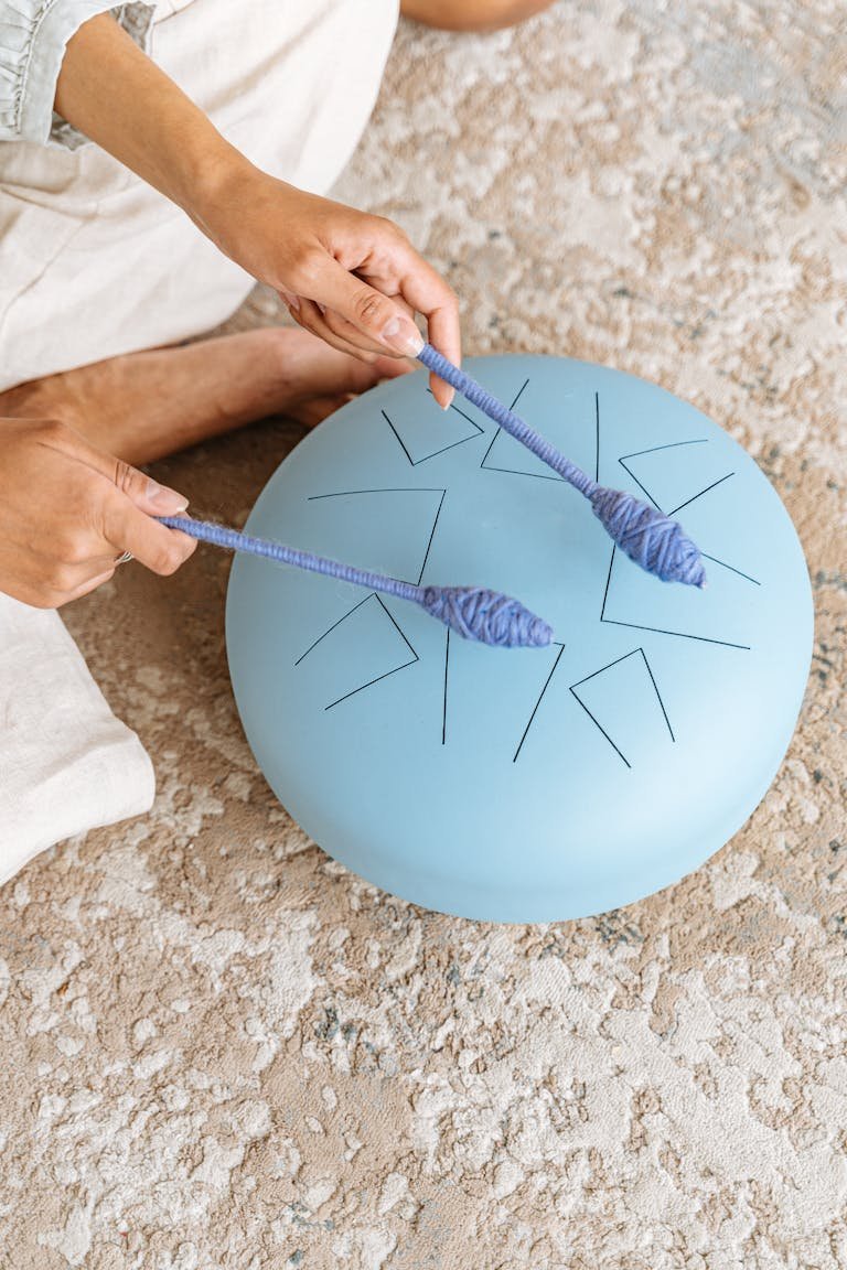 Close-up of hands playing a blue steel tongue drum with mallets on a textured carpet.