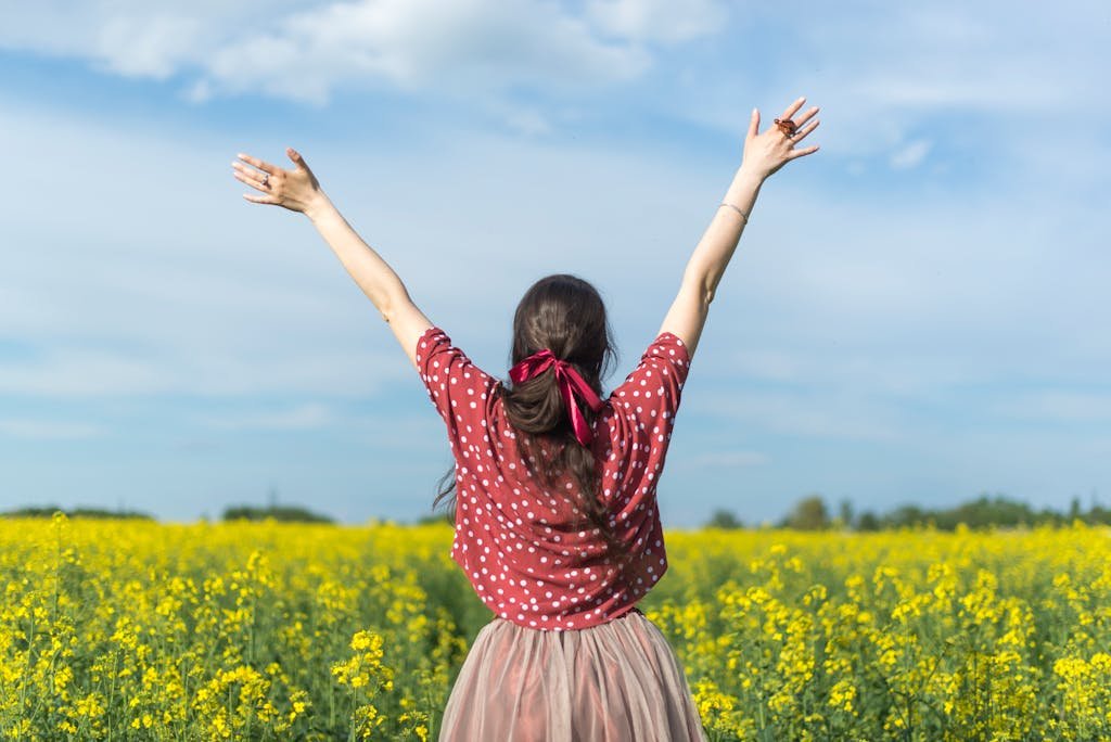 A woman in a polka dot shirt raising her arms in a bright yellow flower field, expressing freedom and joy.